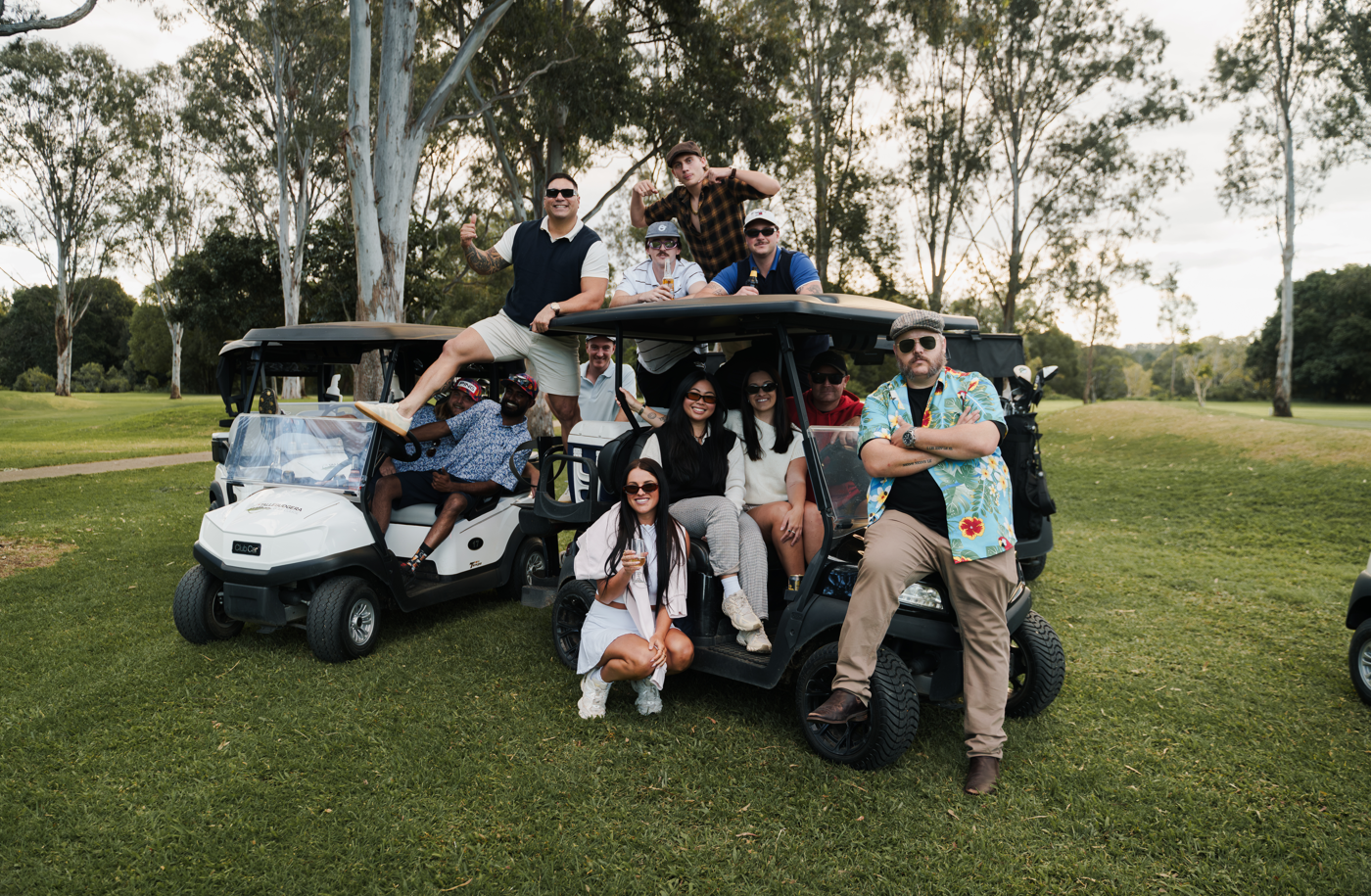 A group of people posing with three golf carts on a grassy golf course. One person is sitting on the roof of the central cart, others are seated inside, and one person is crouching in front.