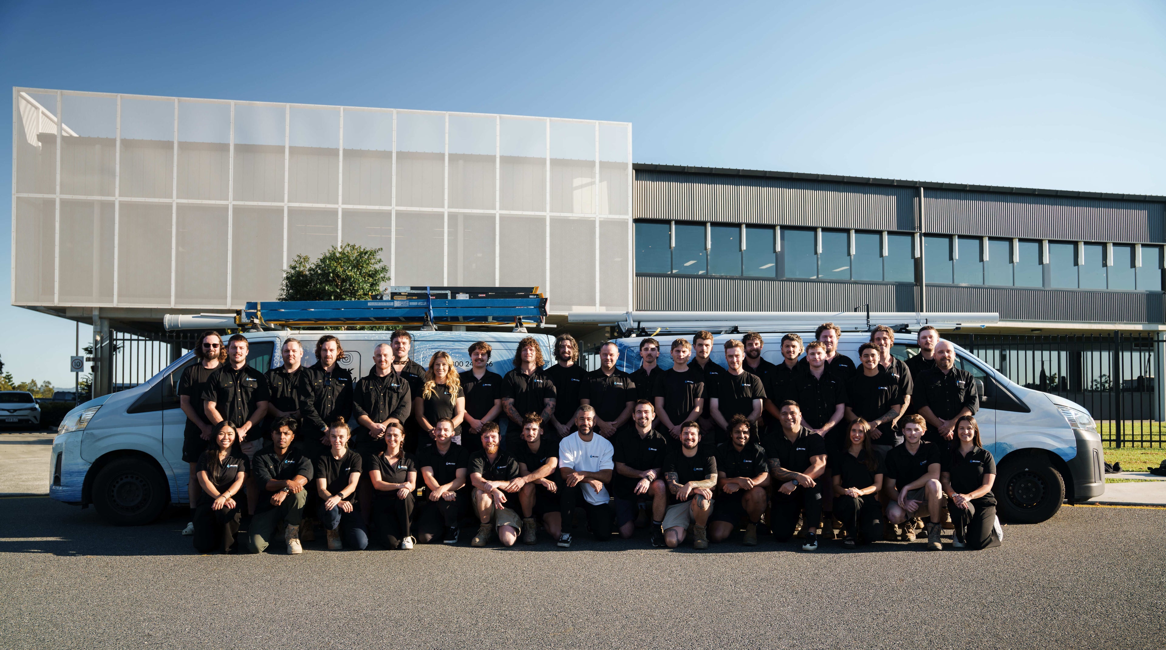 Shark Group Employees posing outdoors in front of a modern building and a white van, with many wearing matching black tops. This is a group photo in a real environment, so the subject type is classified as Lifestyle.